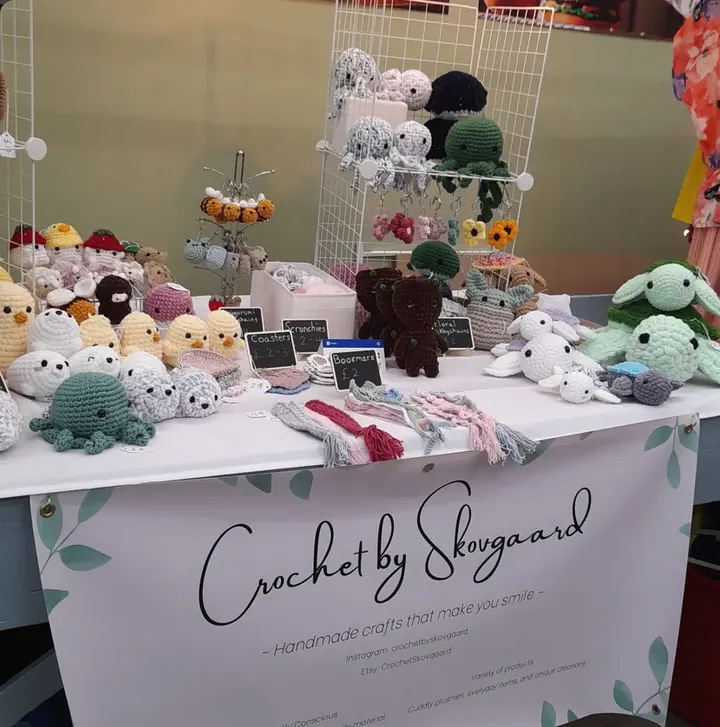 A market stall table of crocheted plushies, keyrings and coasters, with a Crochet by Skovgaard banner hanging in front